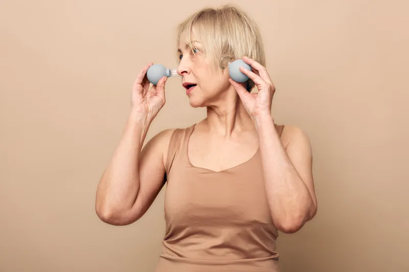 Mature woman in beige tank top holds two gray massage balls near face, looking sideways. Concept for self-care, relaxation, wellness, and facial muscle exercise at home.