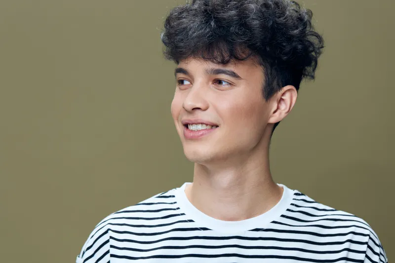 young man with curly dark hair wearing striped t-shirt smiling and looking away against plain olive background, natural light portrait with casual style