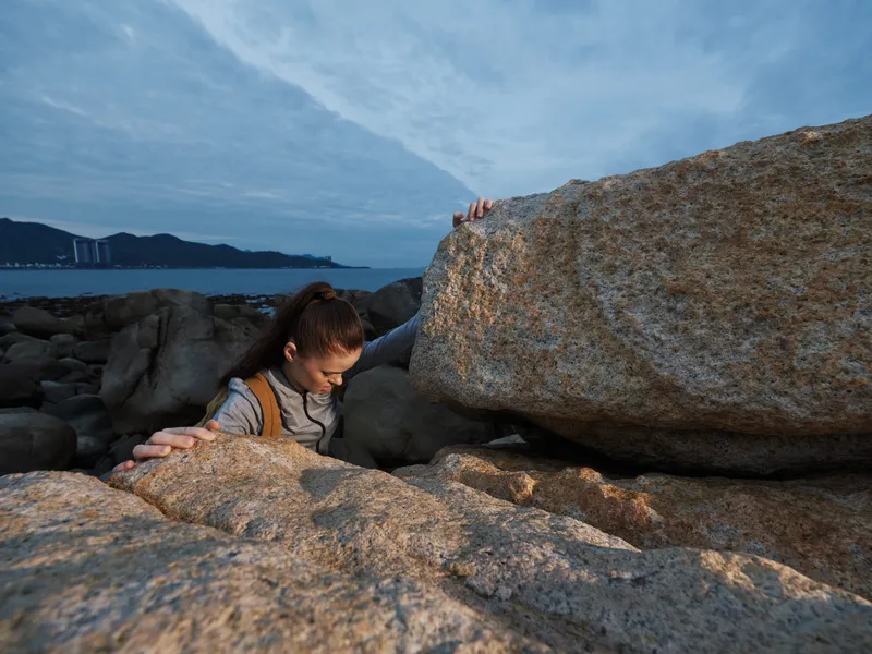 Young woman navigating rocky terrain by the sea, wearing a casual outfit, expressing determination in a natural setting with a serene backdrop