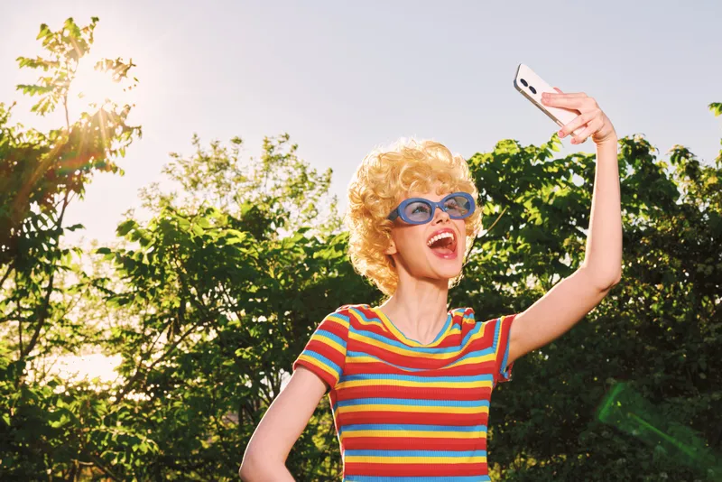 Happy child with curly hair wearing a striped shirt and sunglasses selfies outdoors in bright sun, capturing a playful summer moment with retro film color and cheerful mood.