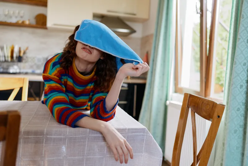 A woman sits at a dining table in a bright kitchen, blindfolded with a blue cloth, smiling softly. A playful home moment exploring senses, mood, and relaxation.