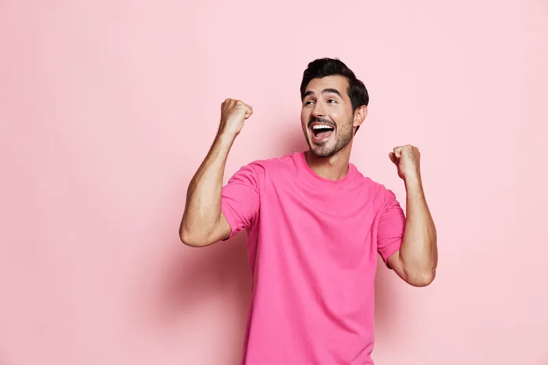 Full body joyful smiling man wearing bright pink t-shirt with enthusiastic expression, raising hands in excitement against solid pastel pink background. Lifestyle concept