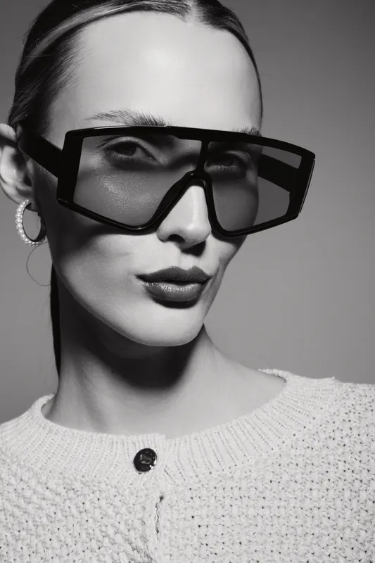 Young white woman with fair skin wears oversized sunglasses and knitted sweater. Studio portrait in black and white.