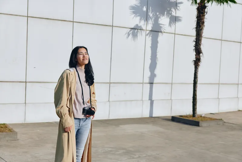 Woman wearing a long trench coat stands outdoors beside a white wall with palm trees, camera hanging, casual fashion and photography vibe for street style and travel, daytime mood, candid urban