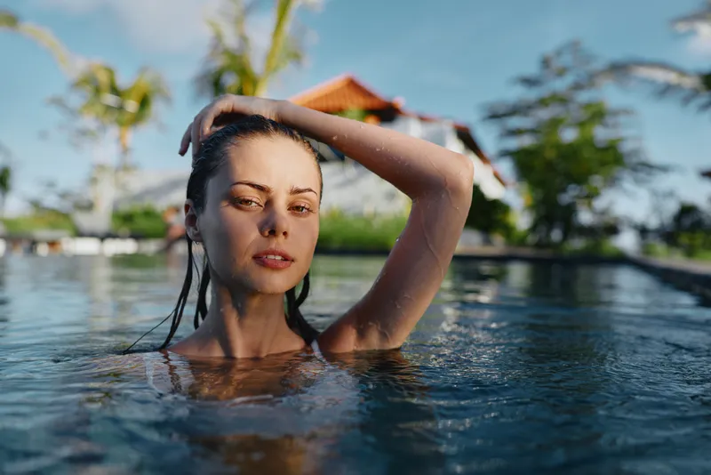 A woman emerges from a turquoise pool, hand in hair, with a tropical resort and palm trees in the background. A lifestyle portrait capturing relaxation, vacation mood, and outdoor leisure.