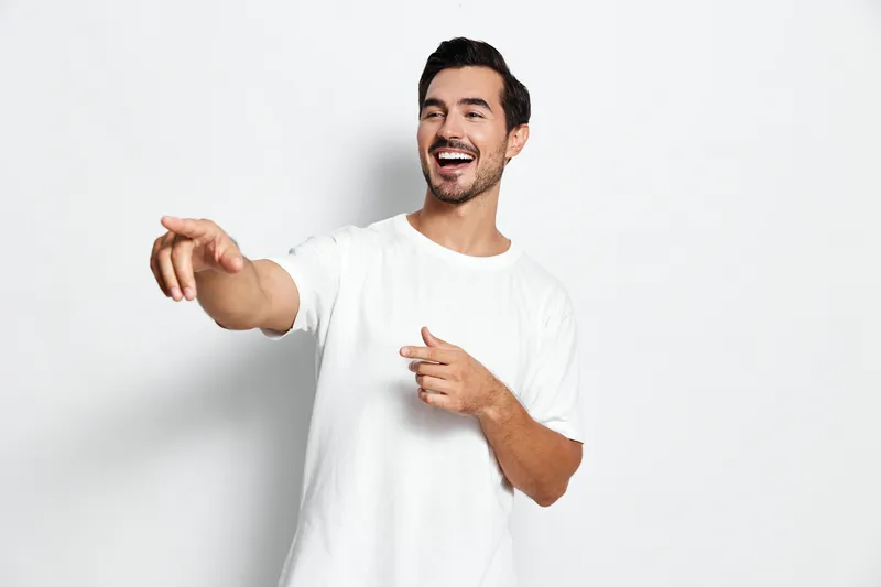 Happy smiling man wearing a white t-shirt gestures while pointing to the side, showcasing a cheerful personality on a light gray background with a warm vibe.
