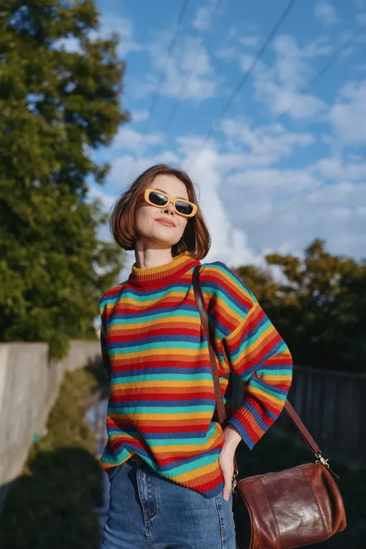 Woman in a rainbow sweater wearing sunglasses and carrying a leather handbag outdoors, casual portrait with blue sky and sunlight, confident pose for street fashion and lifestyle photography.