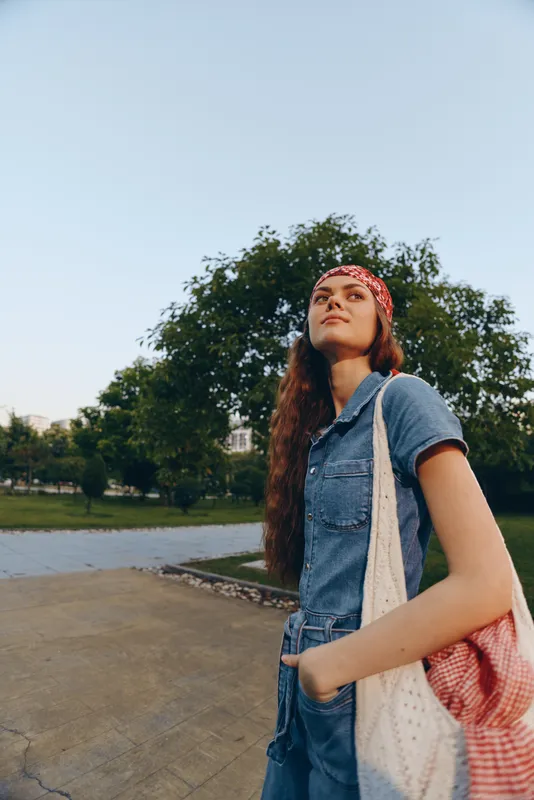 young woman standing outdoors wearing denim jumpsuit and red headscarf holding a tote bag on a sunny day with green trees behind her