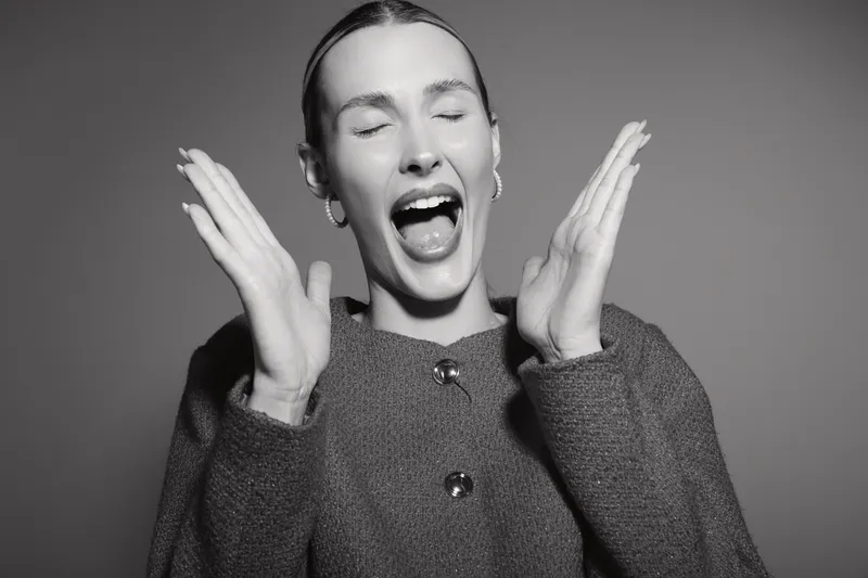 Happy white woman with light skin and tied back hair laughs joyfully with eyes closed and hands raised, wearing a textured jacket in studio. Concept of emotion and happiness.