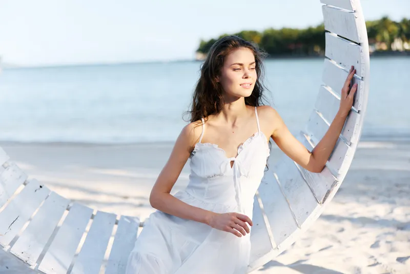 A young woman in a flowing white dress sits on a curved seaside prop, gazing toward the horizon as soft sunlight highlights her calm pose and graceful silhouette on the sand.