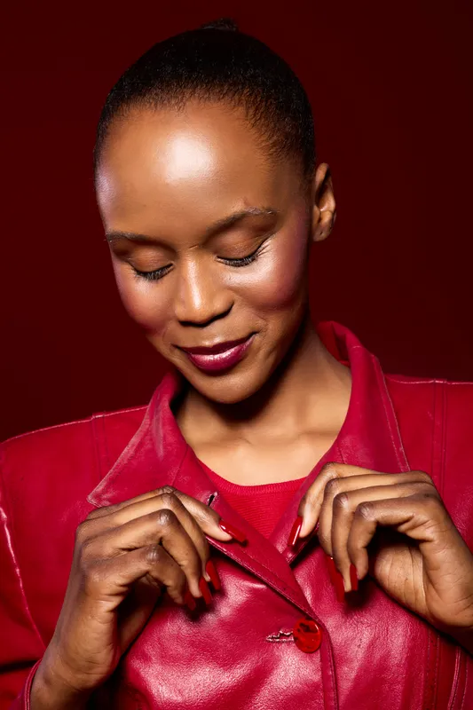 Portrait of a smiling African American woman with dark skin wearing a red leather jacket against a red background. She looks down gently, expressing calm and confidence.