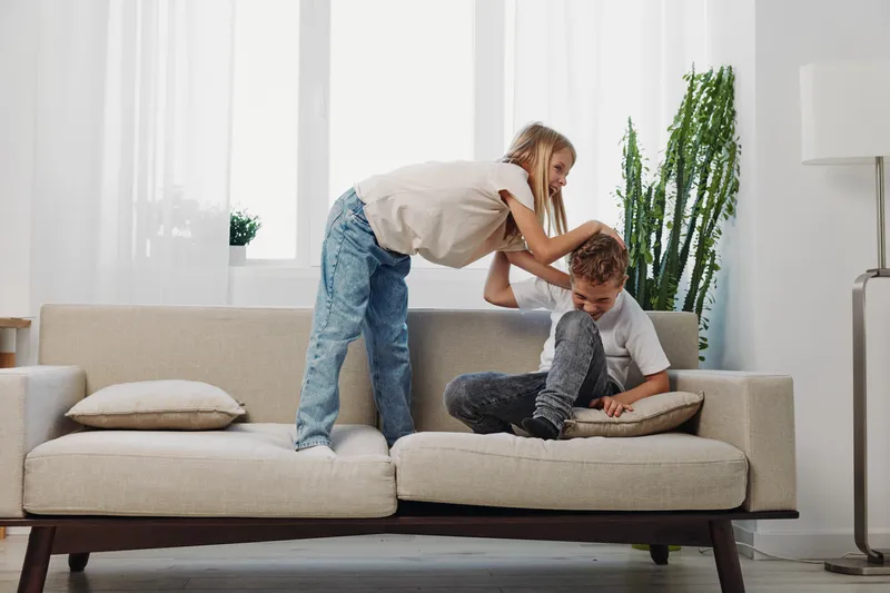 Two children playfully interact on a beige sofa in a bright living room with natural light and indoor plants visible.