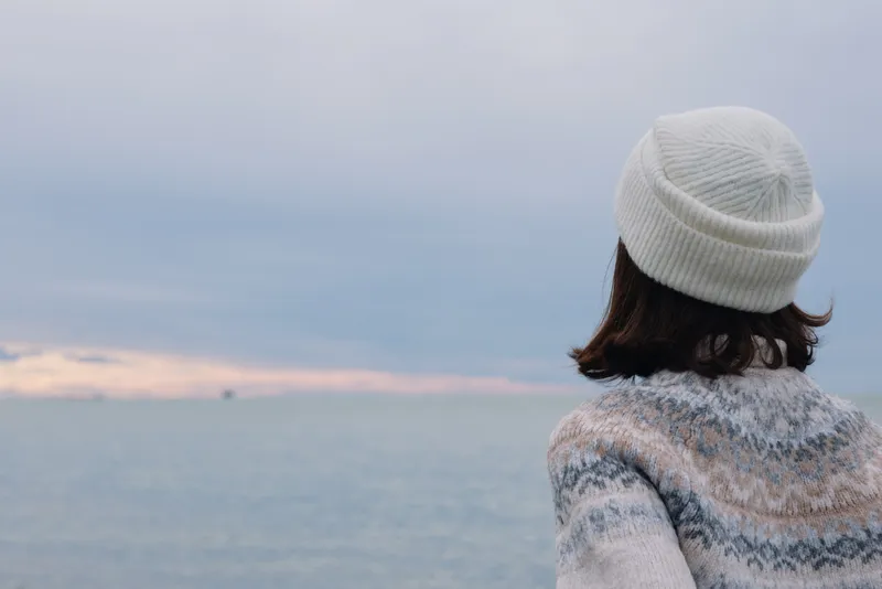 Coast, sea, horizon, woman, winter, knitwear by the water, tranquil scene with soft light, distant clouds and calm mood, scenic overlook capturing quiet reflection and chilly breeze along the