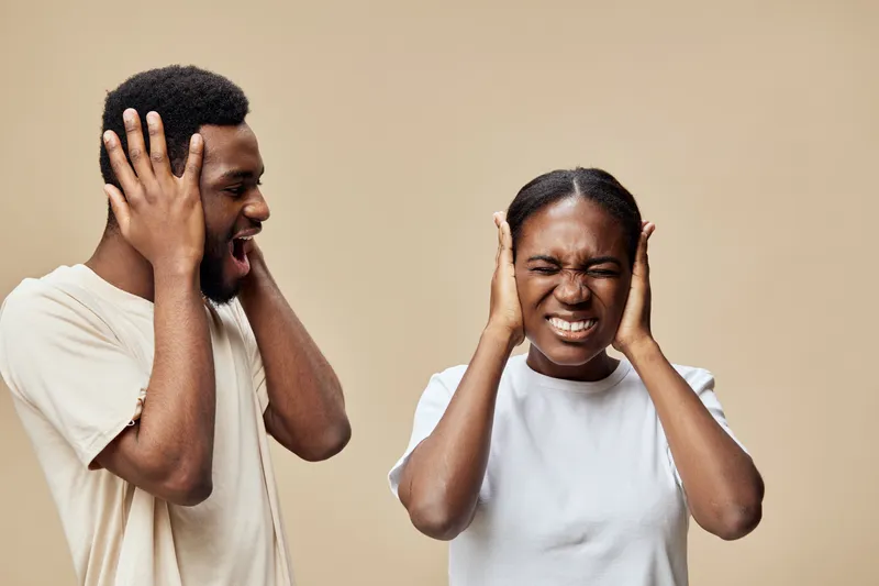 A distressed young African couple expressing frustration and distress, holding their heads in a neutral beige background, capturing raw emotions of stress and anxiety