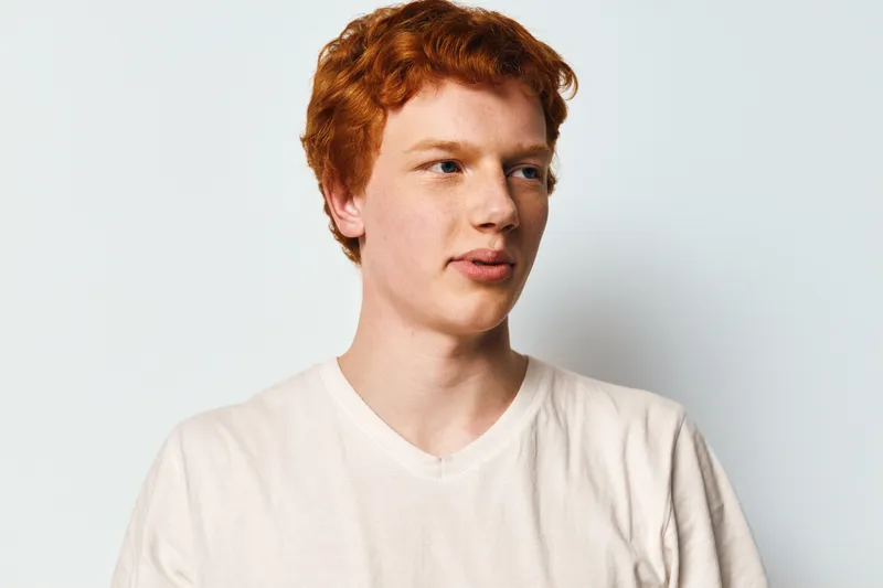 Young man with red hair wearing white t-shirt looking aside with neutral expression on light background. Casual style, portrait, fashion and studio concept with copyspace.