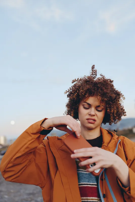 Woman lifestyle outdoor portrait with curly hair, adjusting orange jacket, casual fashion, urban background blurred by bokeh, soft daylight, relaxed confident street mood.