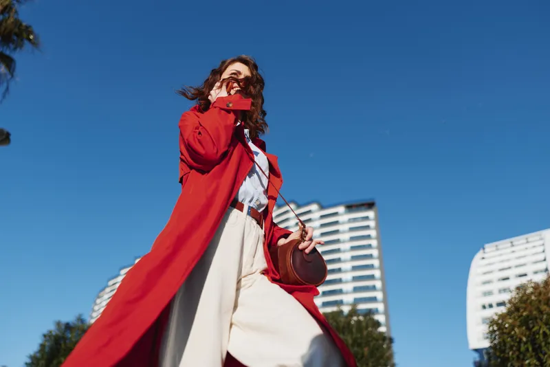 Young woman in a red coat walks outdoors on a bright sunny day, looking up with a confident smile, white trousers fluttering, and a casual crossbody bag swinging.