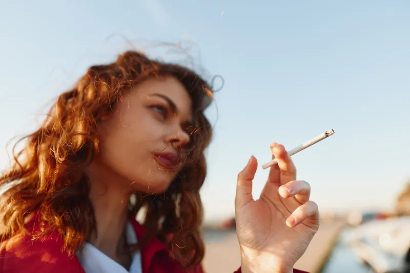 Young woman with curly hair smokes cigarette outdoors in bright daylight with blurred background, thoughtful expression, casual clothing, natural light.