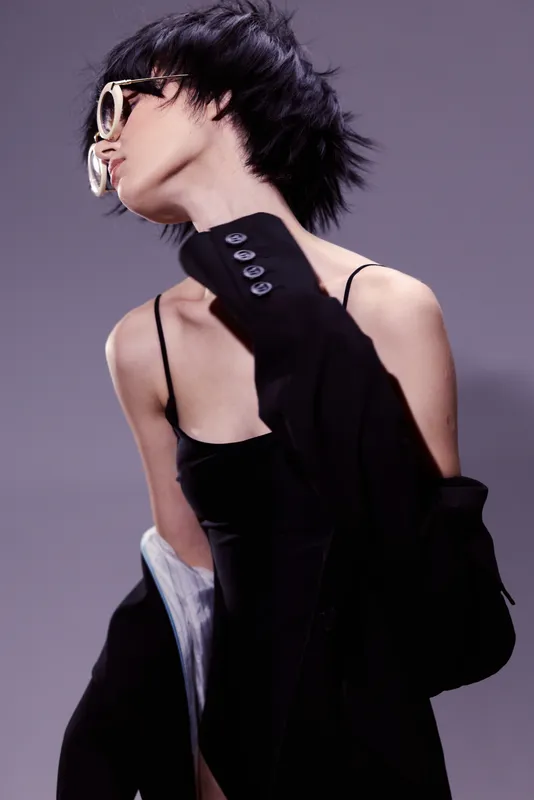 Stylish woman in black dress and oversized jacket posing with sunglasses against gray background