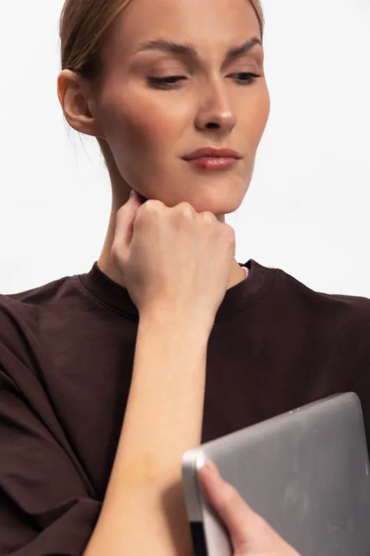 Young woman in dark brown shirt examines skin on her arm holding a tablet with serious expression. Concept of skincare, self-examination, health awareness, and technology use.