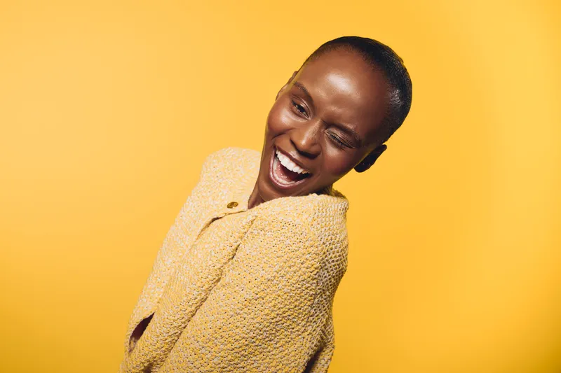 Happy African woman with dark skin winks and smiles wearing yellow sweater. Bright yellow background and casual style create joyful mood. Concept for happiness, confidence, and beauty.
