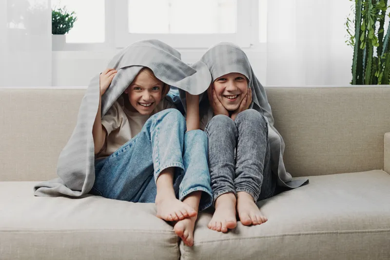 Two happy children sit barefoot on a sofa covered with a blanket, smiling and playing indoors. Cozy home atmosphere with natural light and casual clothing on a bright background.