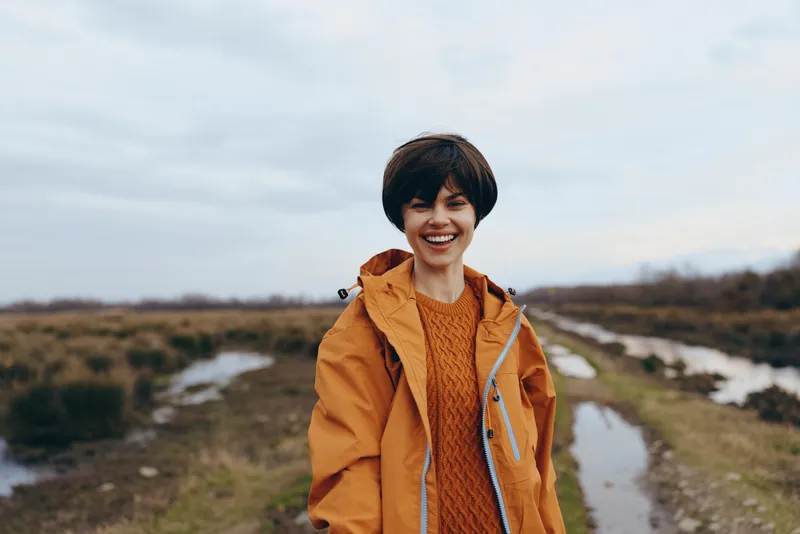 Smiling woman lifestyle outdoor portrait, orange jacket and casual sweater. She stands on a countryside road near grassy fields with small water lines under a cloudy sky.