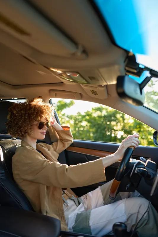 Curly haired woman driving a car, wearing a coat. Close-up interior shot inside a vehicle with sunlight and green trees outside. Lifestyle retro warm sunglasses mood.
