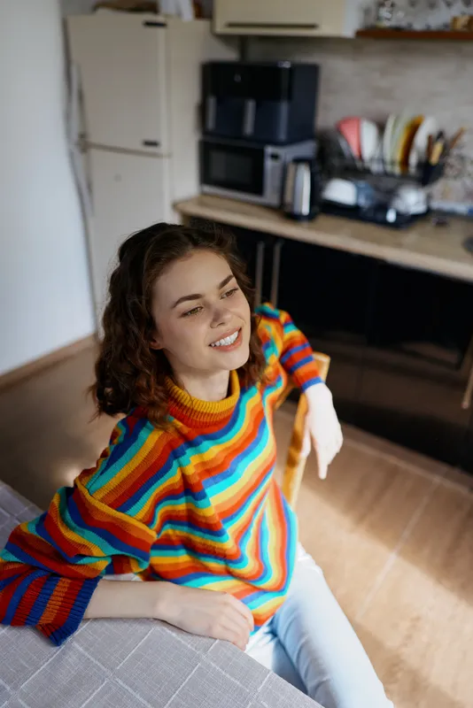 Bright woman indoors, wearing a colorful striped sweater, relaxed smile, cozy kitchen backdrop, natural light, casual lifestyle moment, warm tones, everyday comfort and ease