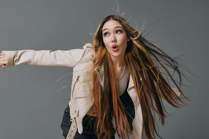 Young woman with long hair wearing beige jacket and white top posing with dramatic expression, hair blowing sideways in motion against gray studio background in casual style.