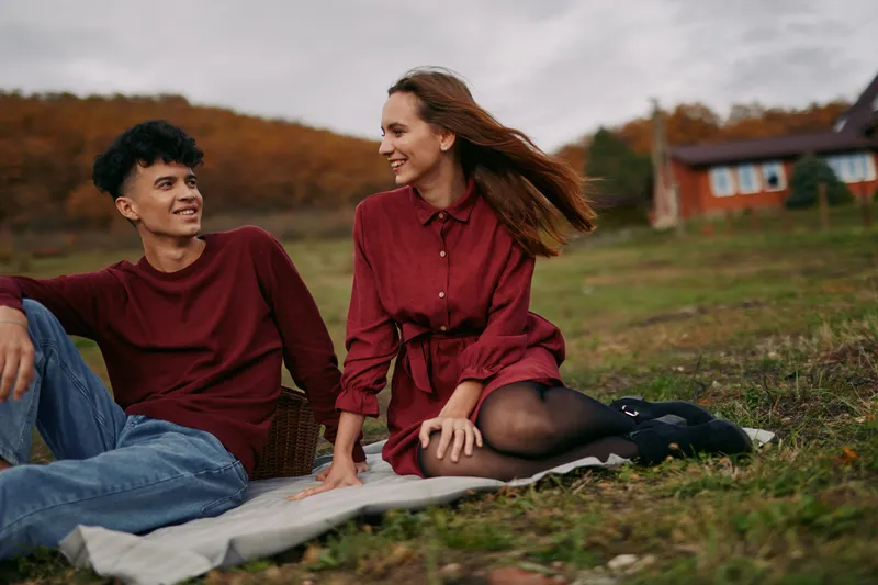 Candid outdoor portrait of a young couple sitting on a blanket, sharing a natural smile and eye contact that conveys authenticity and genuine connection in a relaxed autumn field.