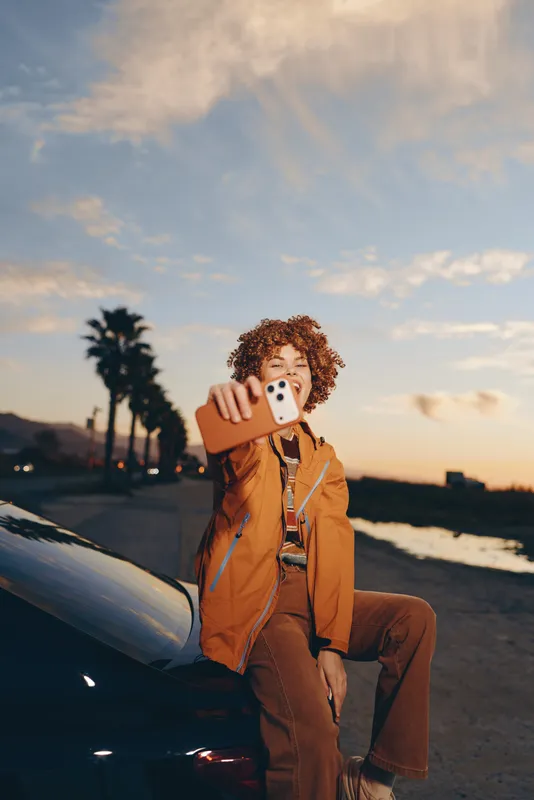 Woman lifestyle at sunset, beach street background, curly haired woman holds a smartphone for a selfie, orange jacket, travel mood, parked car nearby, warm evening light, soft glow