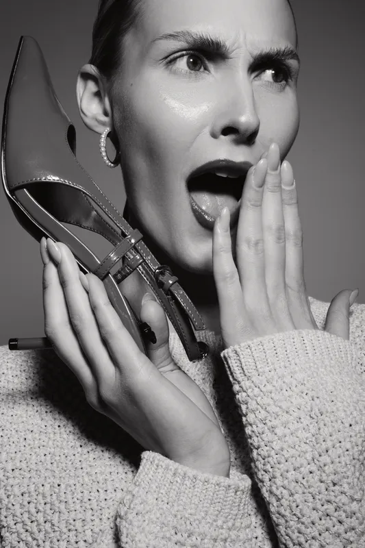 Young white woman with light skin holds shoe near face with open mouth expression on black background.