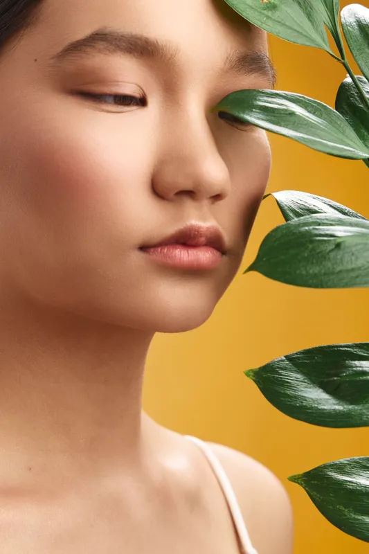 Asian woman poses in a soft studio light with lush green leaves and a warm yellow background, conveying calm beauty, focus, and natural elegance.
