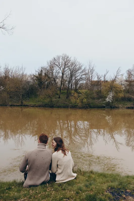 Cheerful couple outdoors, seated by a calm lake, sharing a quiet moment together, autumn colors around them, soft breeze, relaxed posture, casual jackets, reflective water, natural lifestyle scene