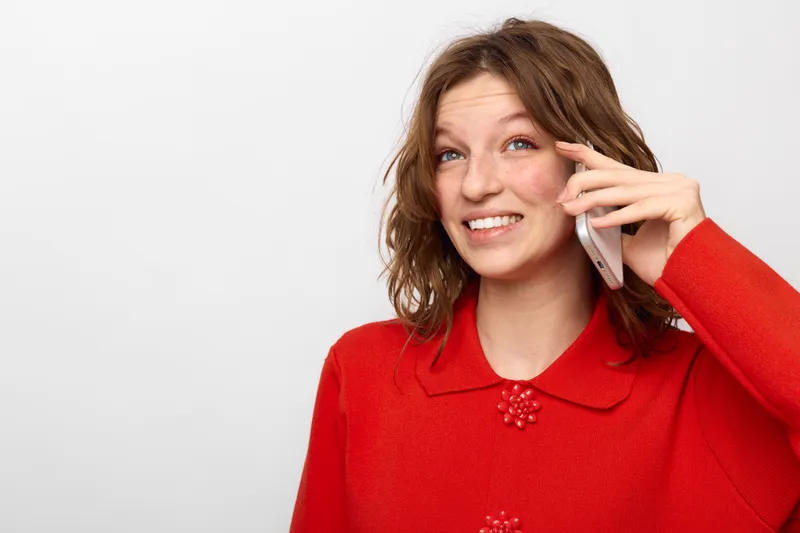 Smiling woman with curly hair wearing red sweater talks on smartphone, looking away. Bright studio background. Concept of communication, technology, and positive emotions.