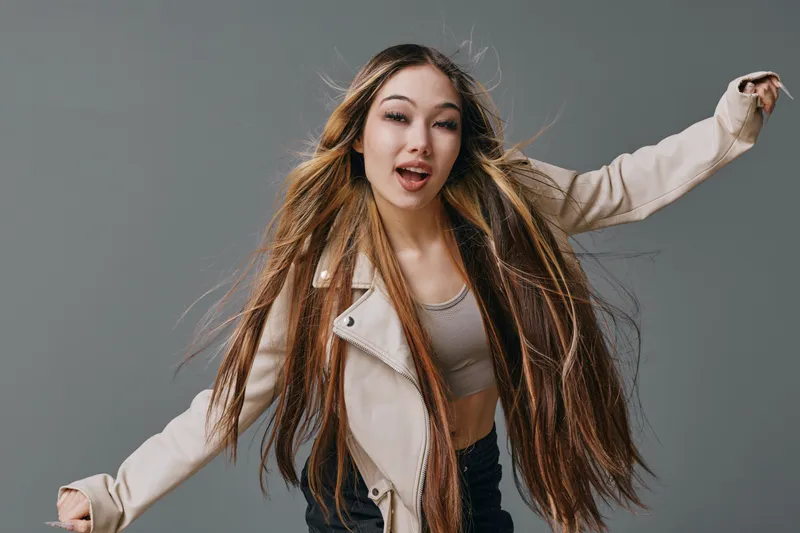 Young woman with long hair wearing jacket and tank top dancing in studio with grey background for beauty, style, youth and fashion expression in motion and energy.
