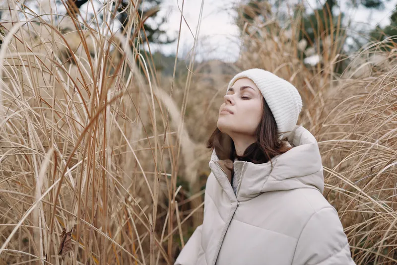 Woman in a warm coat stands among tall dried grasses, eyes closed, savoring a serene winter outdoor nature moment with soft light.
