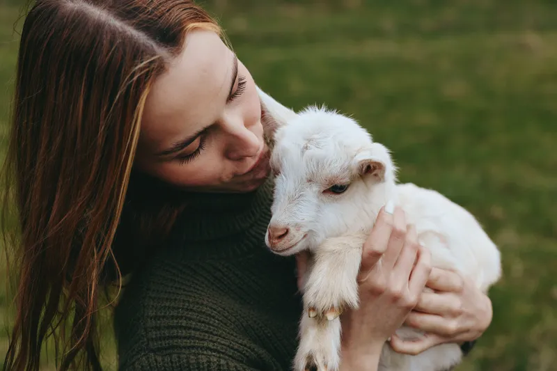 Woman embracing a white goat kid outdoors, tender bonding moment. Natural daylight, grassy field, calm rural lifestyle, affection and trust between human and animal.