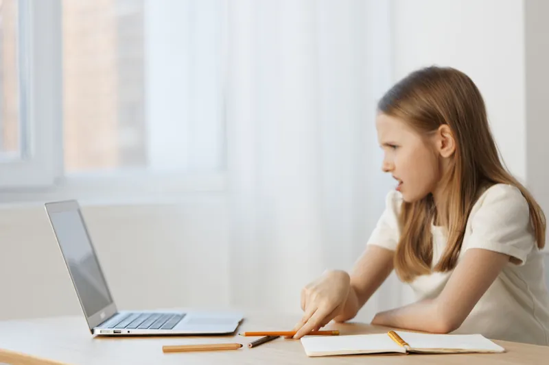 Young girl sitting at a desk, focused on a laptop, expressing concentration while studying at home with natural light flooding in from the window