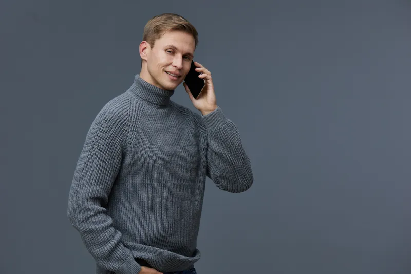 Confident young man in gray sweater talking on mobile phone smiling cheerful casual lifestyle studio portrait. People communication concept