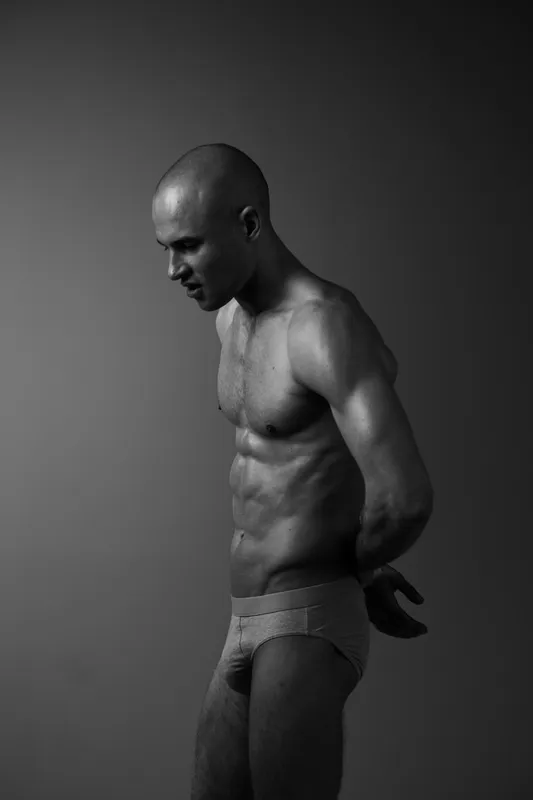 Athletic man posing in a contemplative stance, showcasing defined muscles and a strong silhouette in a striking black and white composition.