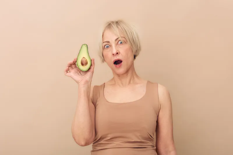 Surprised mature woman with short blonde hair holds half of avocado near face on beige background. Concept for healthy eating, natural food, and nutrition awareness in lifestyle.