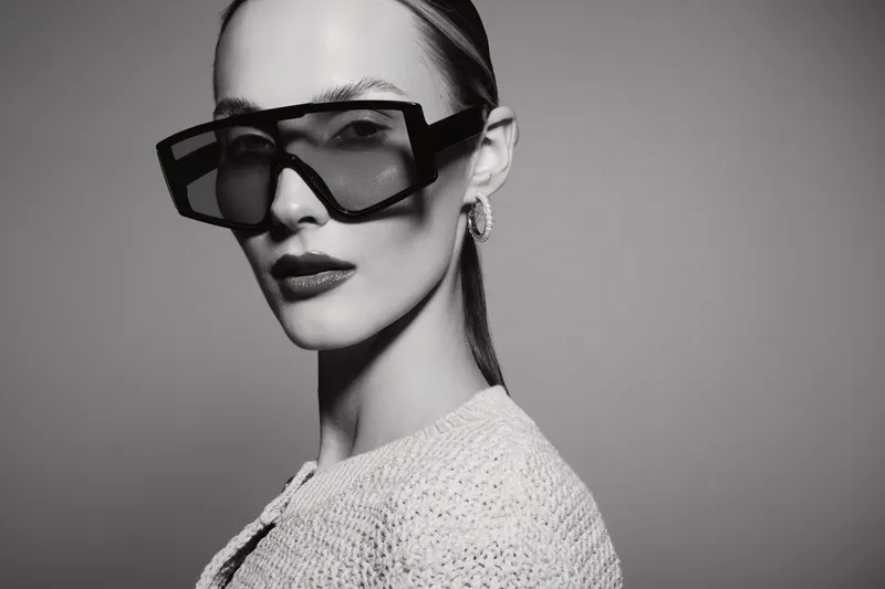 Elegant caucasian woman with pale skin wearing large square sunglasses and earrings looks confidently in monochrome studio. Fashion and style concept.