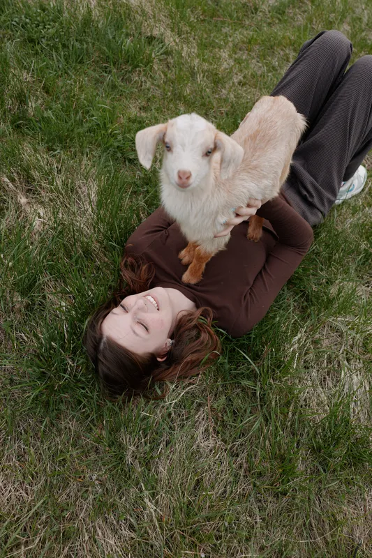 Cheerful woman outdoors, lying in grass with a playful goat kid on her chest, sunlit moment, relaxed mood, natural colors, countryside scenery, gentle breeze, candid lifestyle portrait