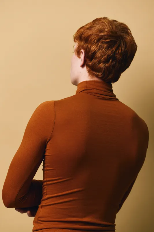 Young man with red hair and fair skin wearing brown turtleneck standing with folded arms against beige background in studio portrait from back.