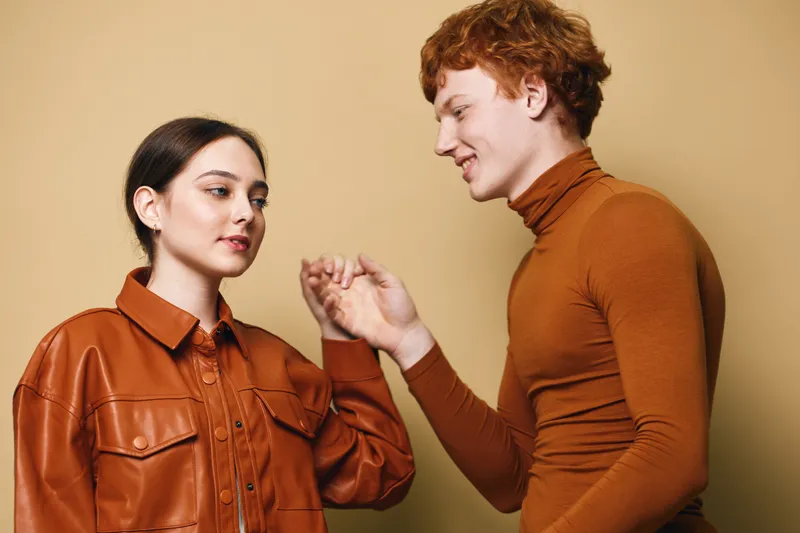 Couple dancing indoors with stylish autumn clothes, young man and woman smiling and holding hands against beige background in casual fashion portrait.