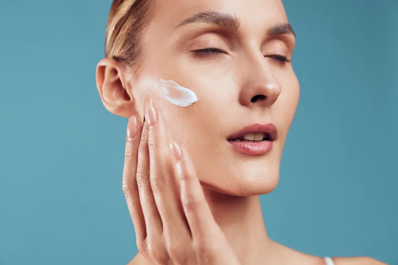 Close-up of woman applying white face cream on cheek with closed eyes. Skincare routine concept with soft natural light and blue background creating calm and fresh atmosphere.