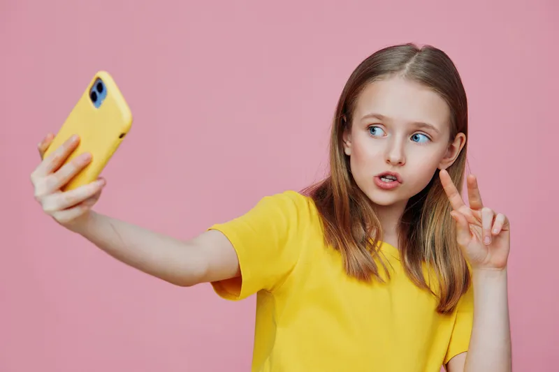 Young girl taking a selfie with a smartphone in a yellow shirt against a pink background, showcasing playful emotions and youthful energy
