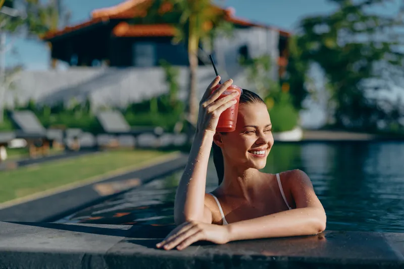 Confident woman enjoys a sunny poolside moment, sipping a pink smoothie, leaning on the edge in a swimsuit, relaxed smile, casual lifestyle, tropical vibe, daytime leisure, cheerful mood, serene